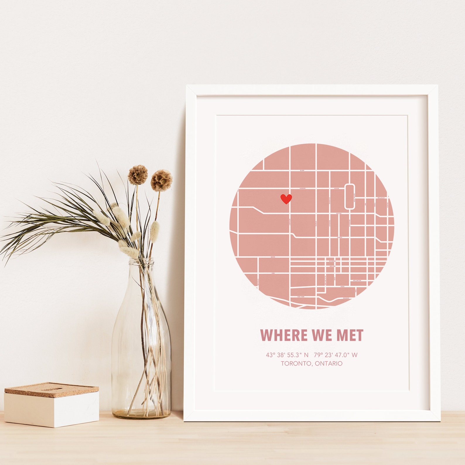 Image of map in a pink circle with type below that says Where We Met and coordinates on the next line. Frame is placed on a wood surface beside a decorative plant in a clear bottle.