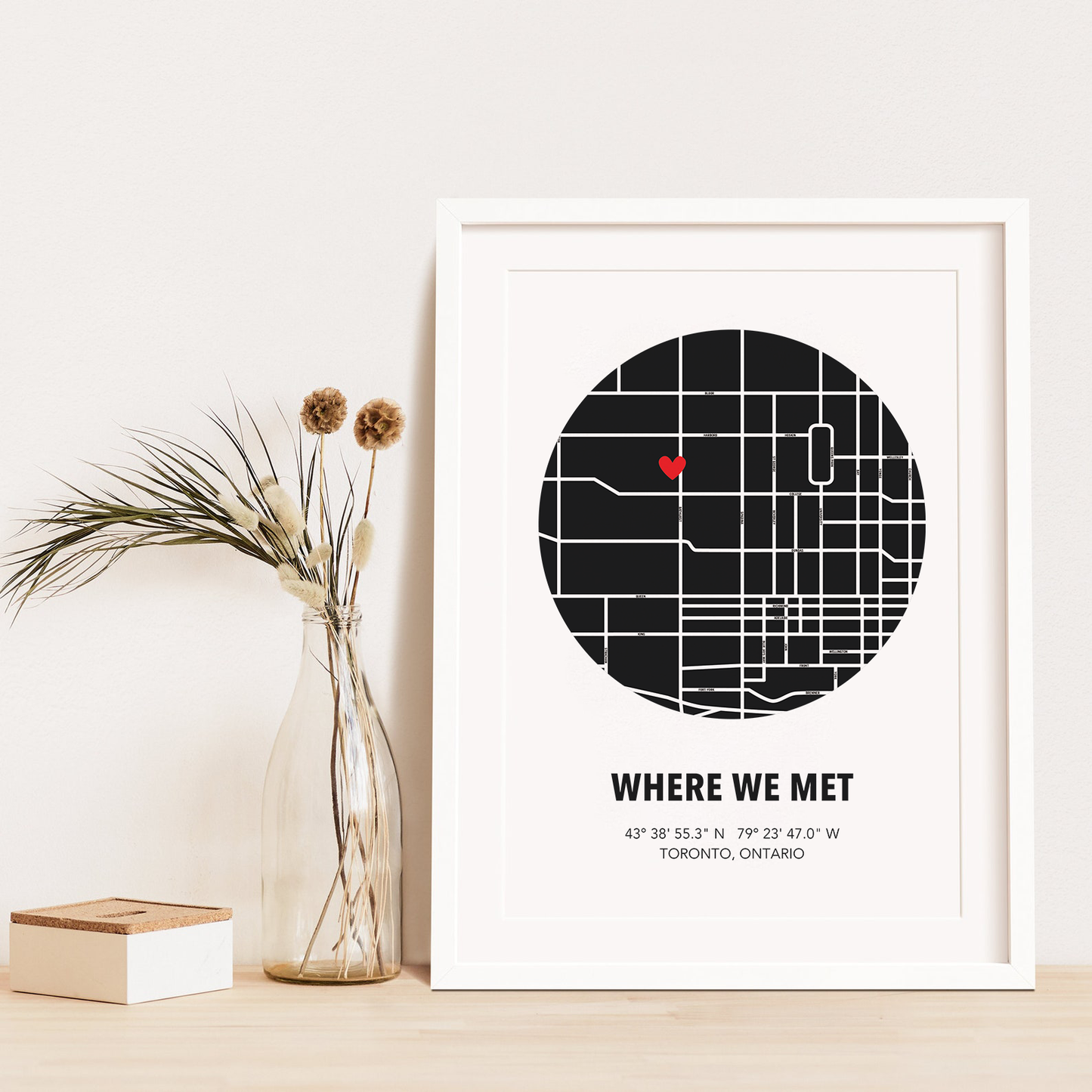 Image of map in a black circle with type below that says Where We Met and coordinates on the next line. Frame is placed on a wood surface beside a decorative plant in a clear bottle.