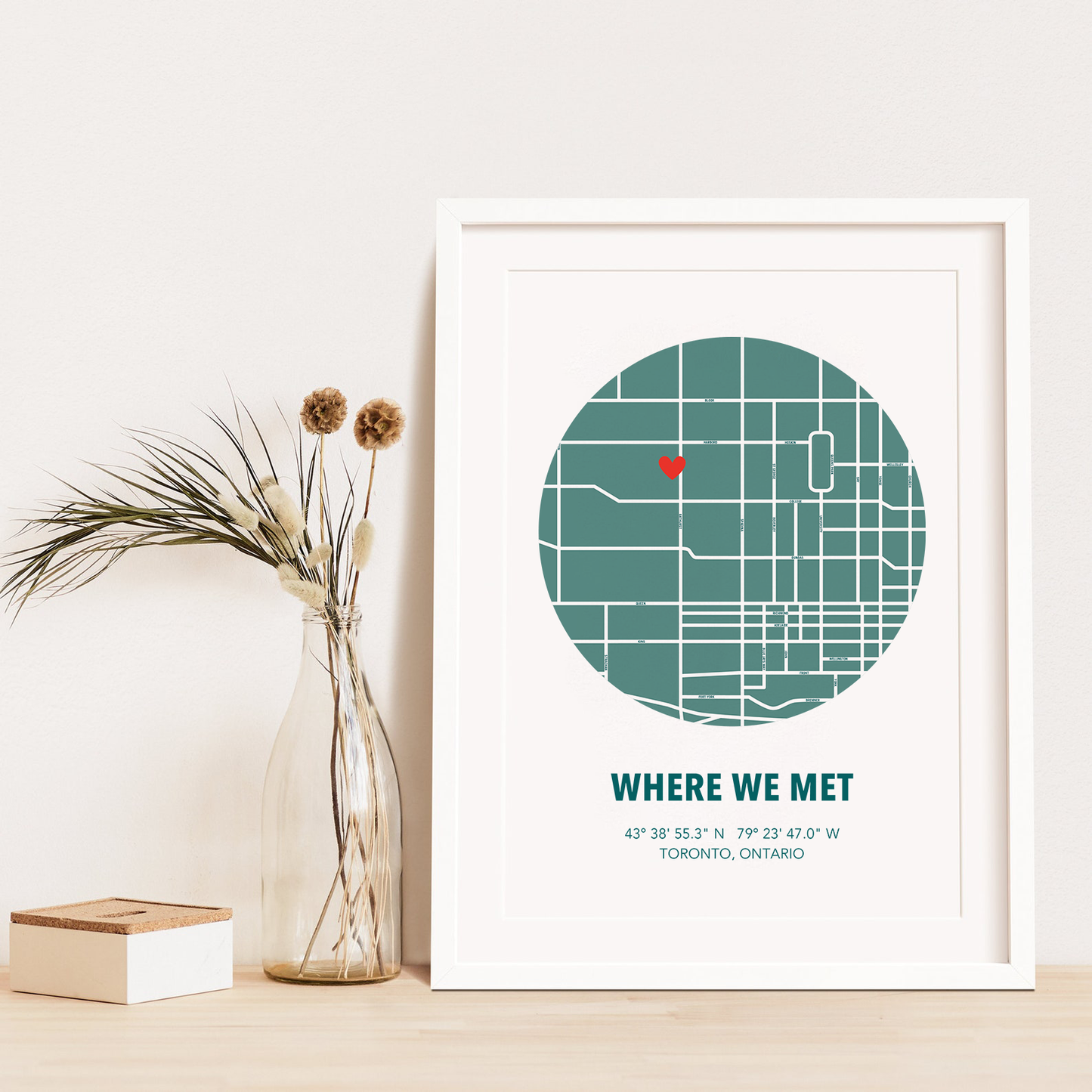 Image of map in a green circle with type below that says Where We Met and coordinates on the next line. Frame is placed on a wood surface beside a decorative plant in a clear bottle.