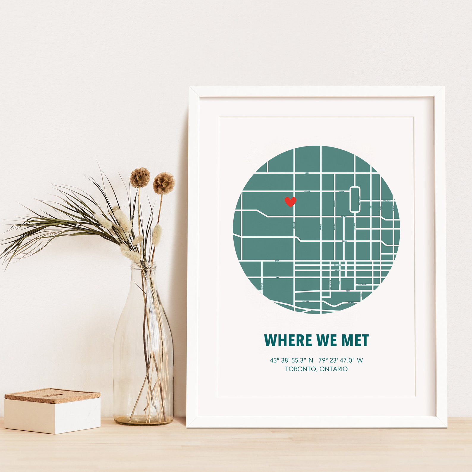Image of map in a green circle with type below that says Where We Met and coordinates on the next line. Frame is placed on a wood surface beside a decorative plant in a clear bottle.