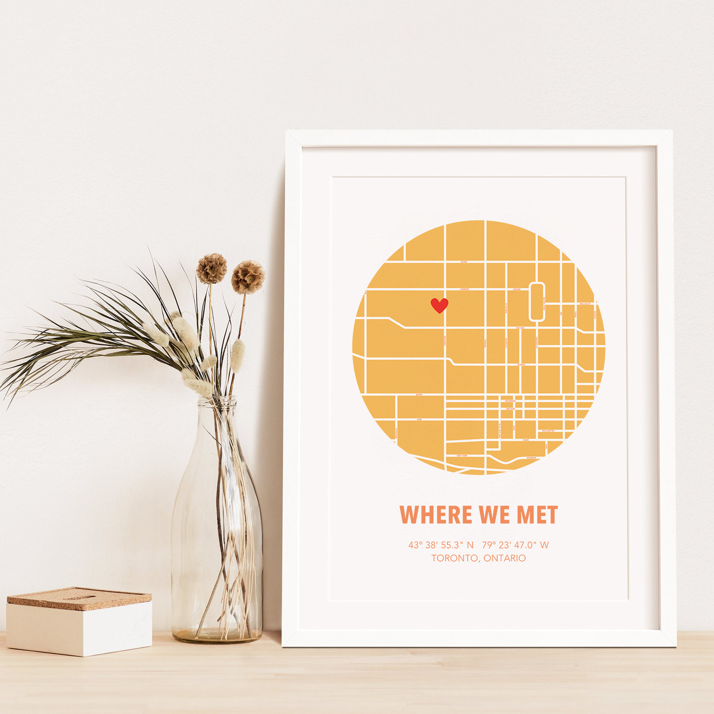 Image of map in a yellow circle with type below that says Where We Met and coordinates on the next line. Frame is placed on a wood surface beside a decorative plant in a clear bottle.
