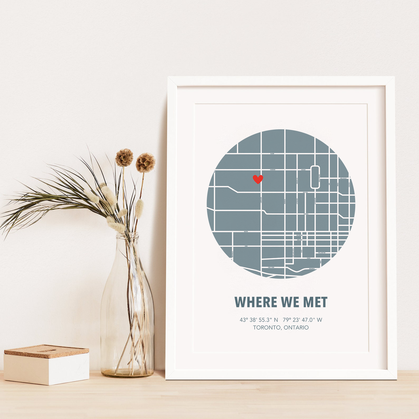Image of map in a blue circle with type below that says Where We Met and coordinates on the next line. Frame is placed on a wood surface beside a decorative plant in a clear bottle.
