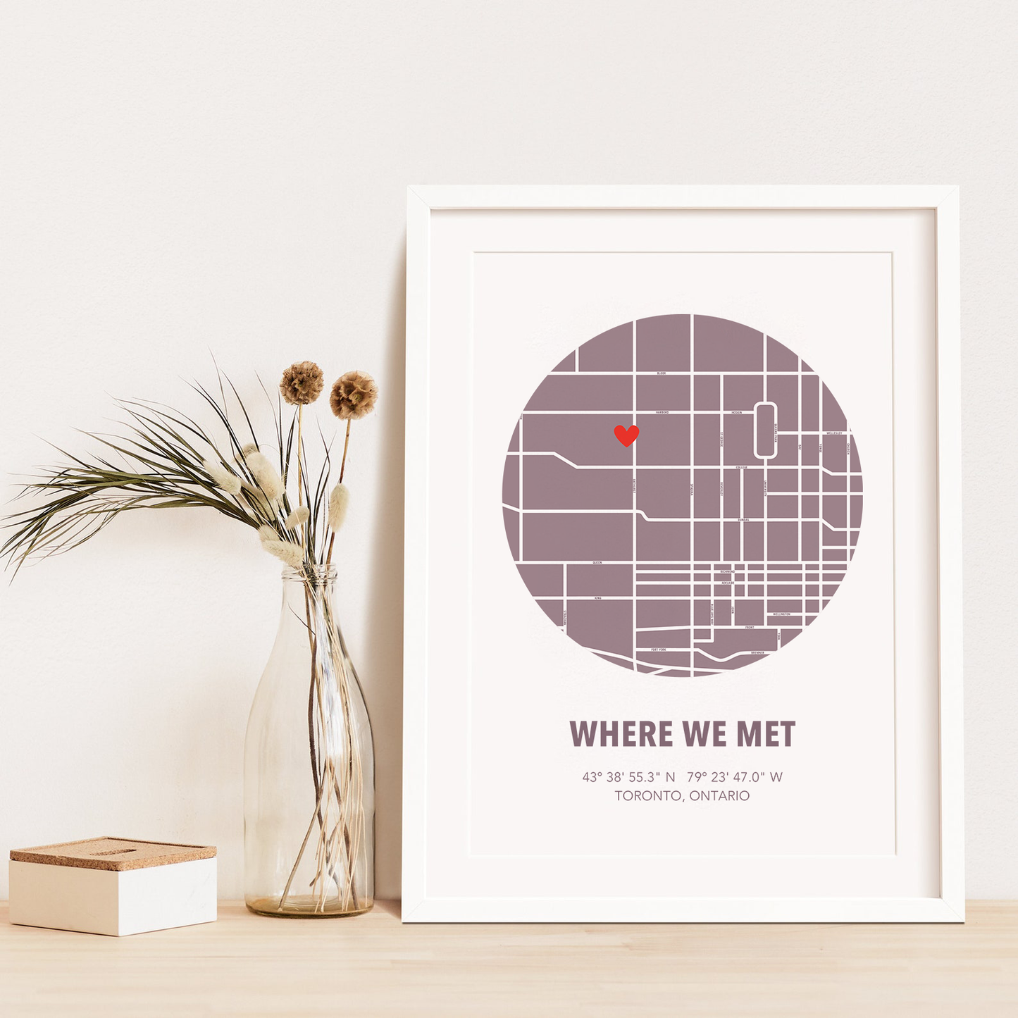 Image of map in a mauve circle with type below that says Where We Met and coordinates on the next line. Frame is placed on a wood surface beside a decorative plant in a clear bottle.