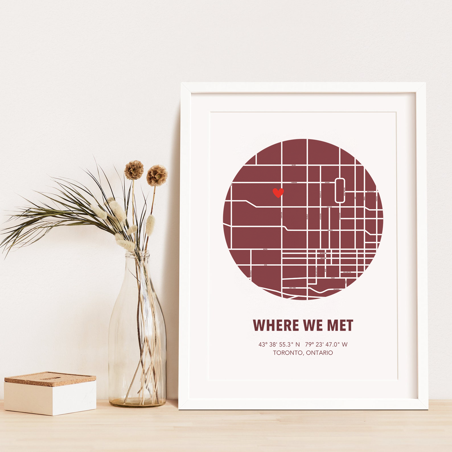 Image of map in a maroon circle with type below that says Where We Met and coordinates on the next line. Frame is placed on a wood surface beside a decorative plant in a clear bottle. 